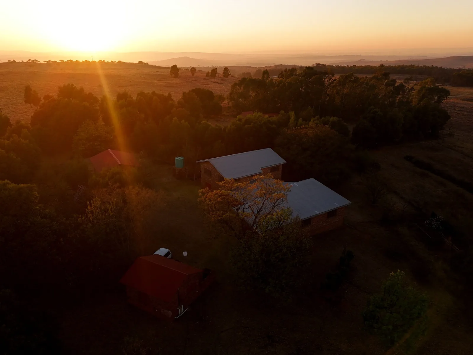 A smallholding homestead photographed from the air at sunrise, the surrounding grassland and hills stretching to the horizon.